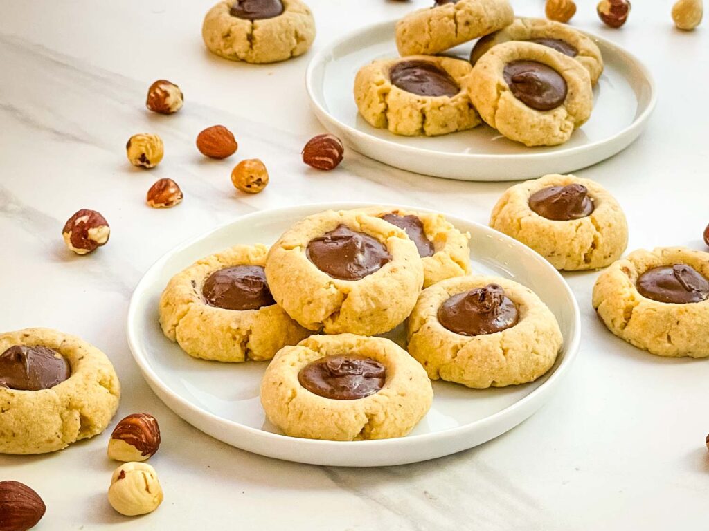 Two small white plates full of Chocolate Hazelnut Thumbprint Cookies with extra cookies and nuts scattered around them.