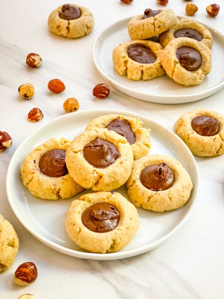 Chocolate Hazelnut Thumbprint Cookies on two small plates on a counter with more cookies and nuts around them.