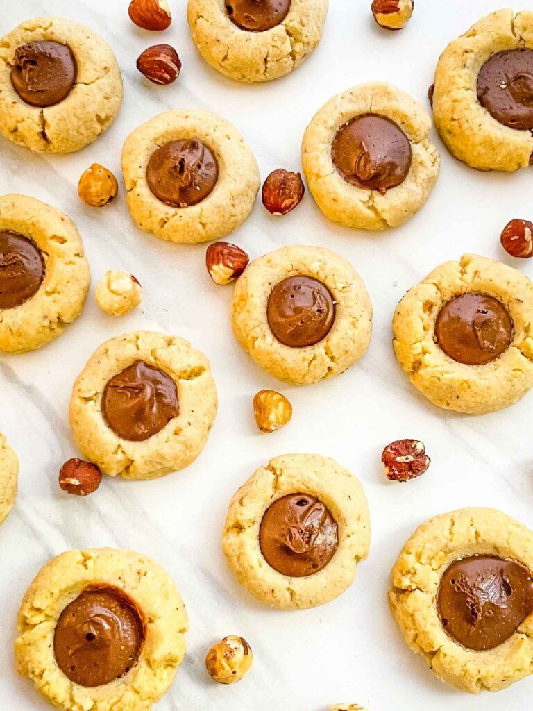 Chocolate Hazelnut Thumbprint Cookies spread out on a counter, looking down on them, surrounded by whole hazelnuts.