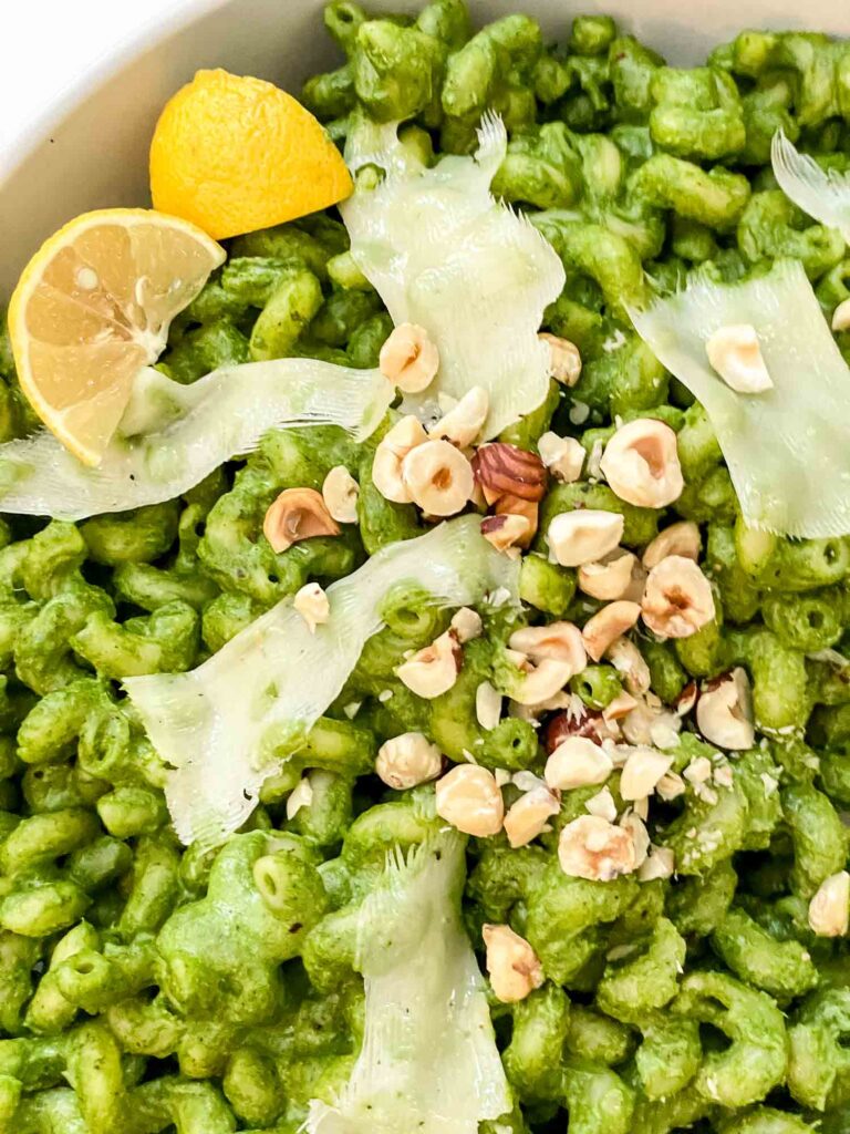 Close view of Hazelnut Spinach Pesto Cavatappi garnished with chopped nuts, lemon wedges, and cheese shards.