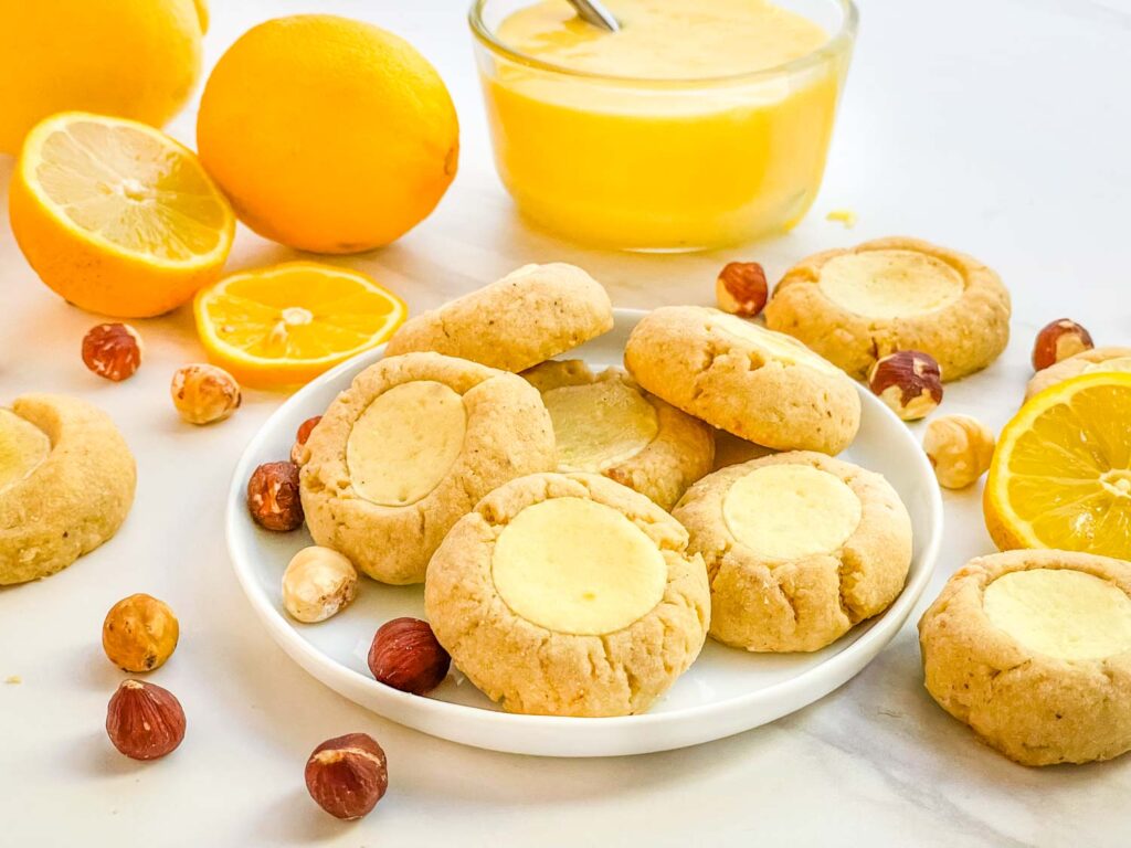 A small white plate with Hazelnut Lemon Curd Thumbprint Cookies surrounded by nuts and lemon.