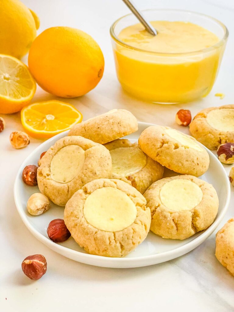 A small pile of Hazelnut Lemon Curd Thumbprint Cookies on a counter with ingredients around them.