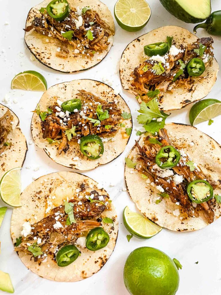 Flat Pulled Pork Carnitas Street Tacos spread out over a white counter with lime wedges.