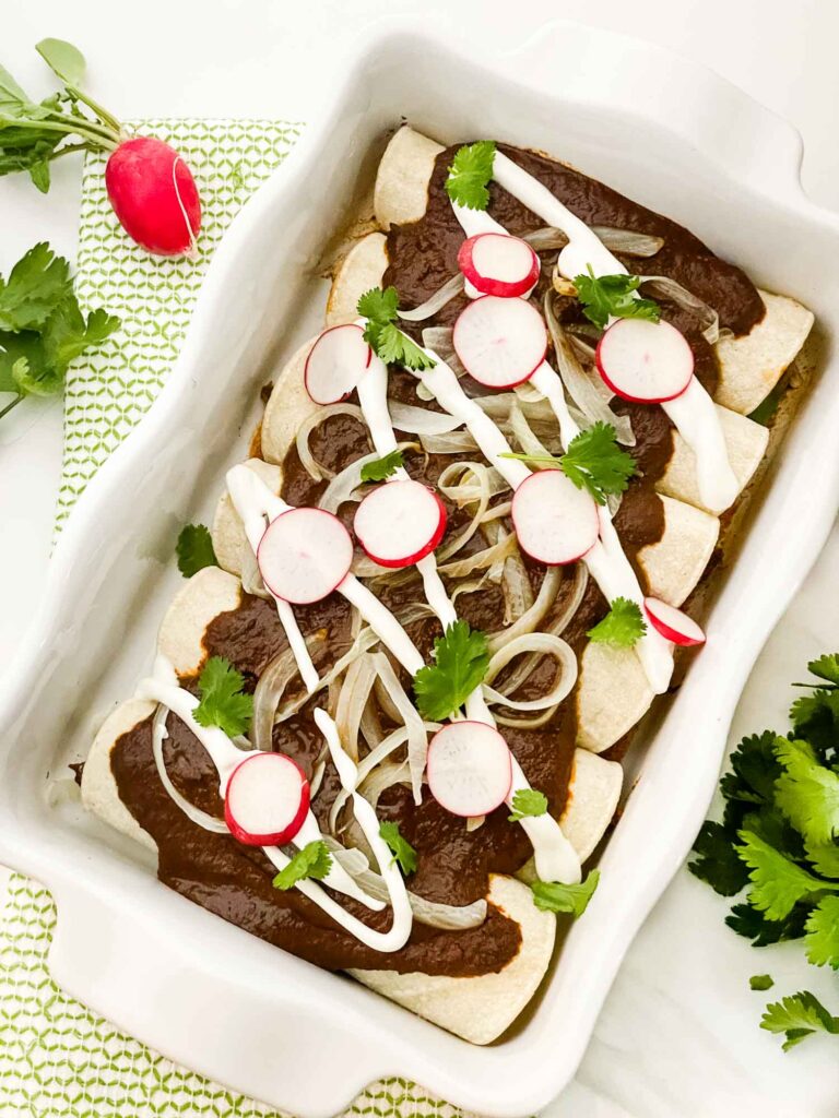 Looking down on a white rectangular dish full of Chicken Mole Enchiladas with toppings.