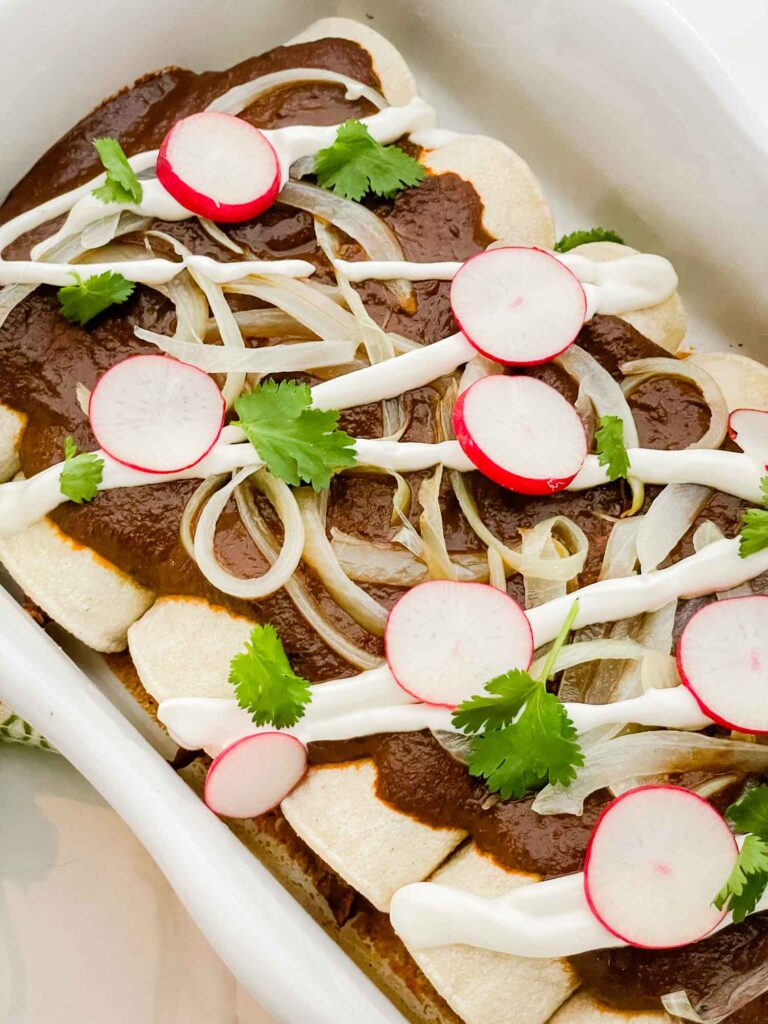 A closer view of Chicken Mole Enchiladas in a white dish with radishes, sour cream, onions, and cilantro.