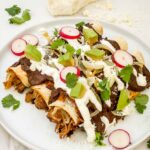 A white plate on a counter with three Chicken Mole Enchiladas and fresh toppings on and around them.