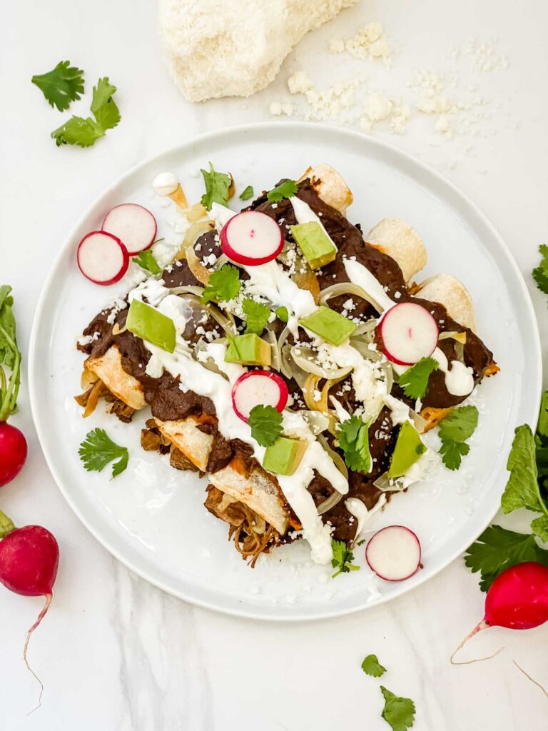 Looking down on three Chicken Mole Enchiladas on a white round plate with radishes, cilantro, avocado, and sour cream on them.
