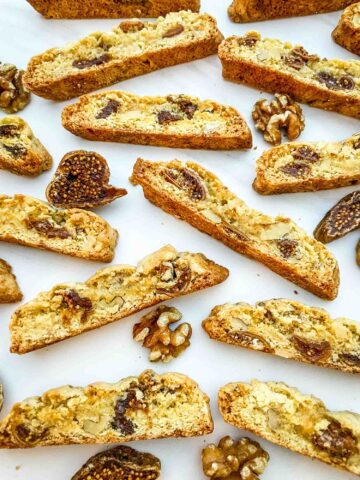 Fig Walnut Biscotti cookies spread out on a counter with nuts and fruit around them.
