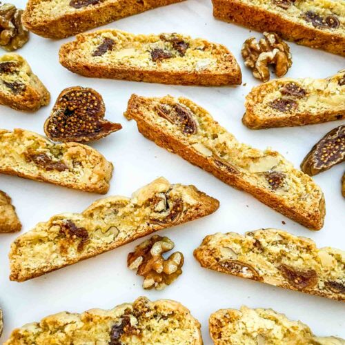 Fig Walnut Biscotti cookies spread out on a counter with nuts and fruit around them.