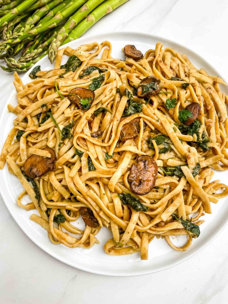 Looking down on a plate of Mushroom Marsala Wine Sauce pasta on a counter with asparagus next to it.