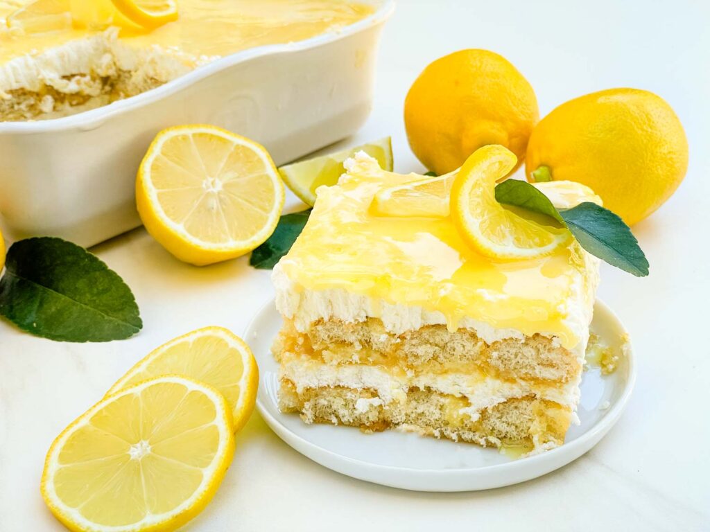 A piece of layered Limoncello Tiramisu on a small plate next to the serving dish and surrounded by fresh lemons.