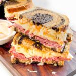 Two halves of the Corned Beef Pastrami Reuben Sandwich Recipe stacked on a wood board.