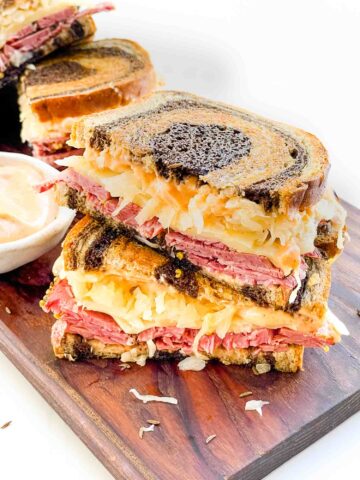 Two halves of the Corned Beef Pastrami Reuben Sandwich Recipe stacked on a wood board.