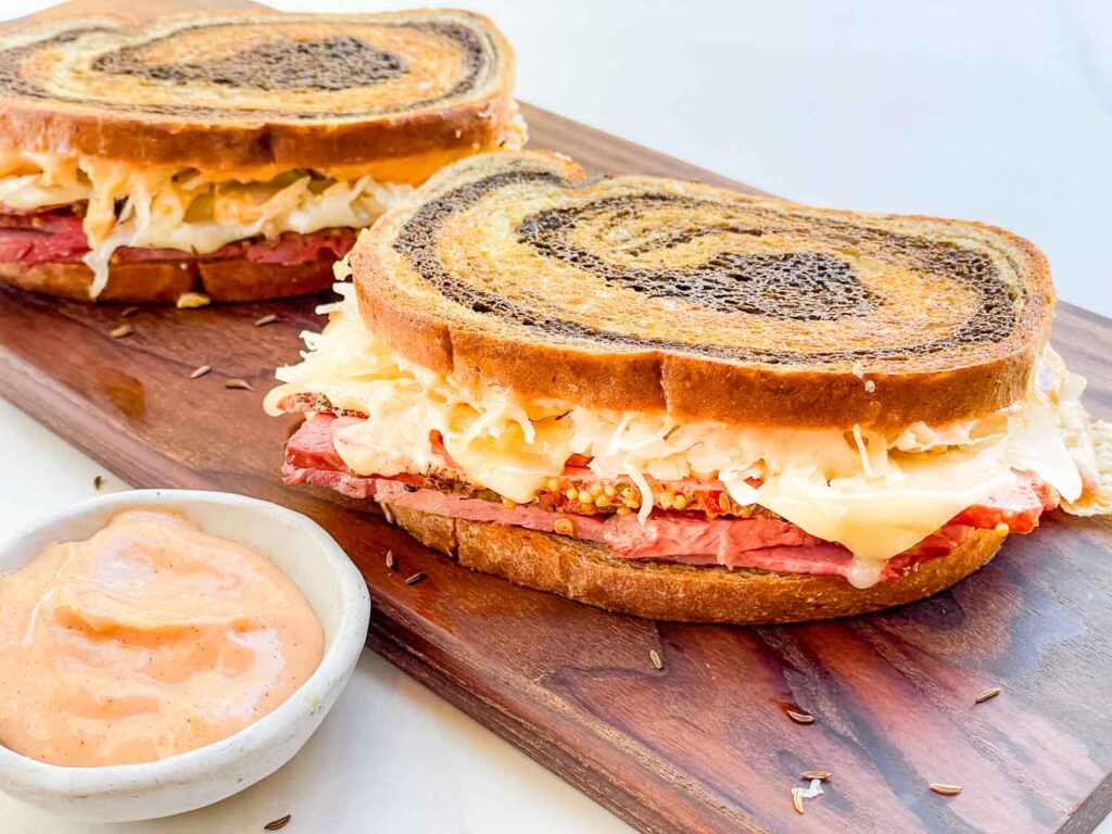 Two whole corned beef Pastrami Reuben Sandwiches on a wood board.