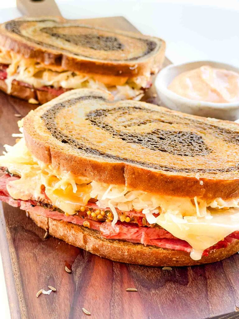 Two whole corned beef Pastrami Reuben Sandwiches sitting on a wood board.