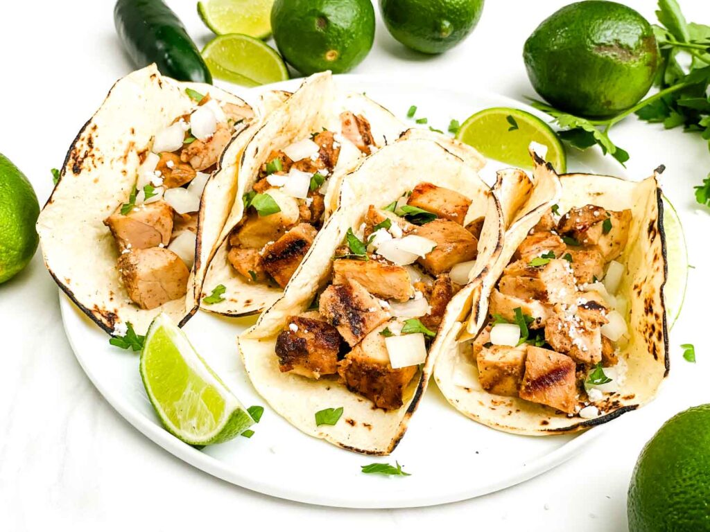 Four Chicken Street Tacos on a white plate on a counter surrounded by limes.