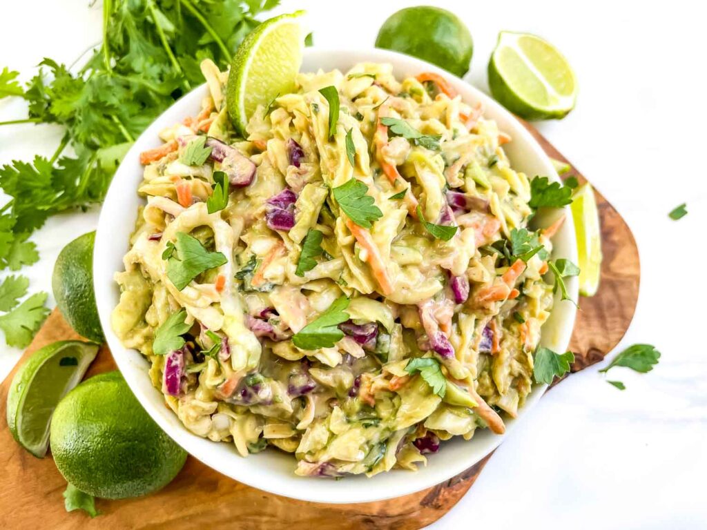 Looking down on a bowl of Creamy Avocado Coleslaw (without Mayo) on a wood board and surrounded by limes.