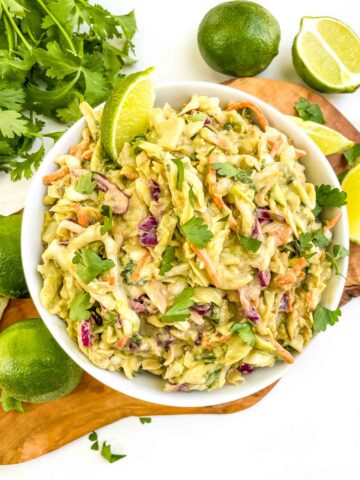 Looking down at a white bowl of Creamy Avocado Coleslaw (without Mayo) on a wood board on a counter.