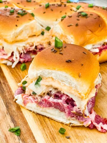 A single slider in front of other Corned Beef Reuben Sliders on a cutting board.