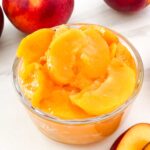 A small glass bowl full of Easy Peach Dessert Sauce surrounded by fresh peaches.