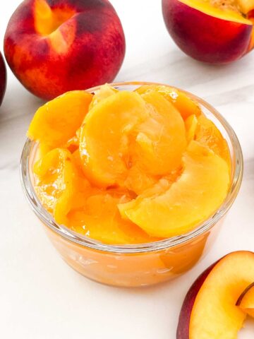 A small glass bowl full of Easy Peach Dessert Sauce surrounded by fresh peaches.