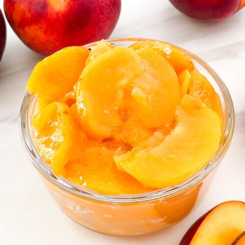 A small glass bowl full of Easy Peach Dessert Sauce surrounded by fresh peaches.