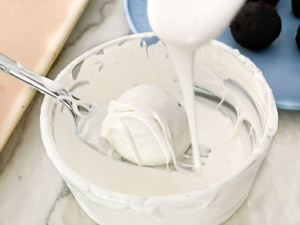 A spoon drizzling strips of white candy melts over a coated cake ball to make mummy bandages.