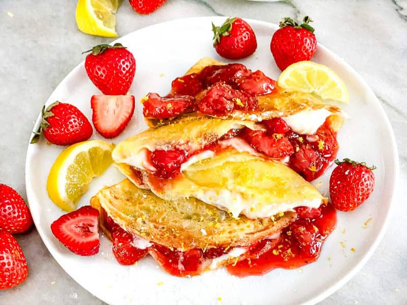 A close view of Lemon Strawberry Crepes on a plate with fresh fruit around them.