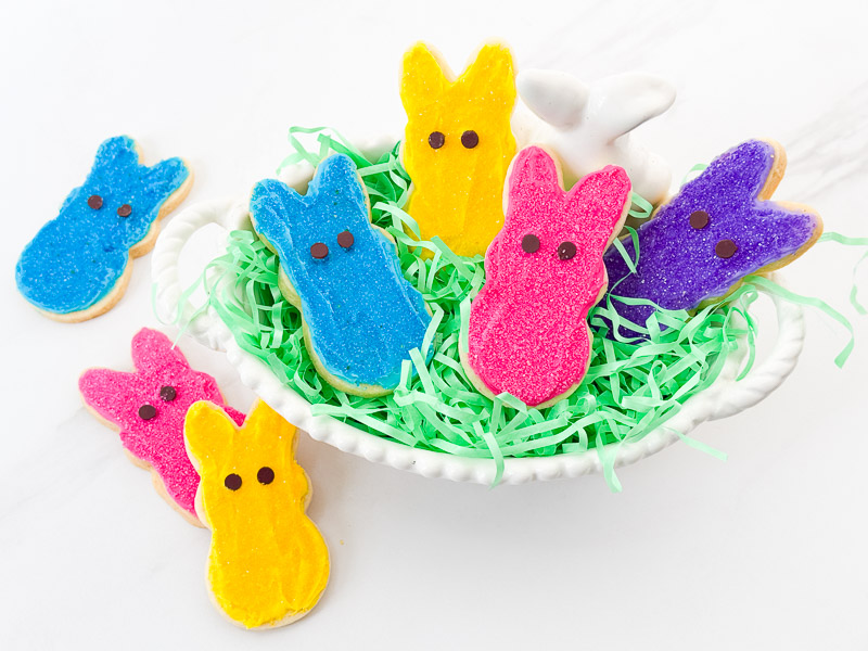 Peeps Bunny Easter Sugar Cookies in a white basket dish with green Easter grass on a counter with some extra cookies around it.