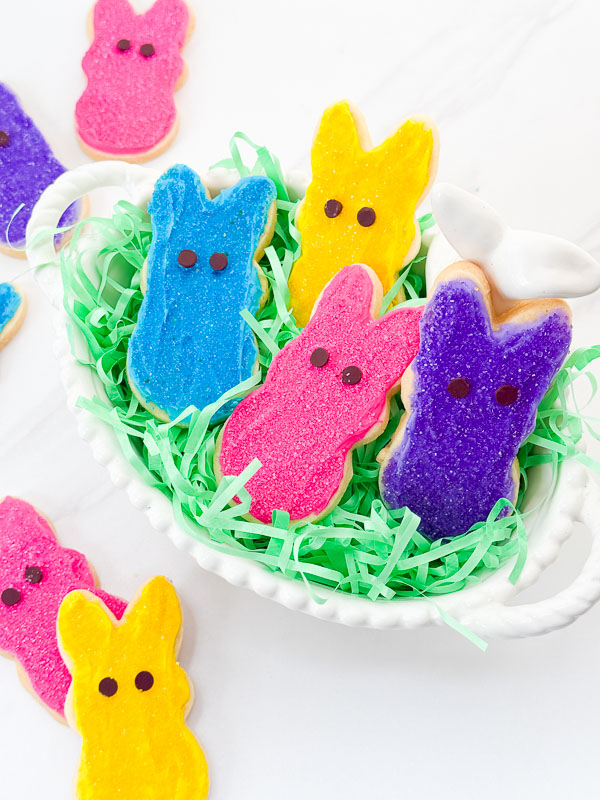 Looking down on Peeps Bunny Easter Sugar Cookies in a white ceramic basket with green Easter grass on a counter.