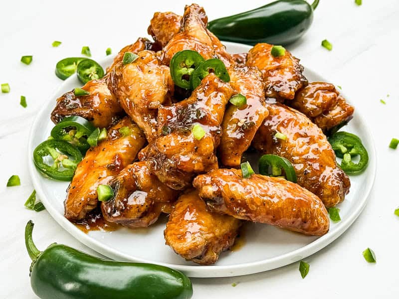 A pile of Peach Jalapeno Chicken Wings on a plate surrounded by peppers.