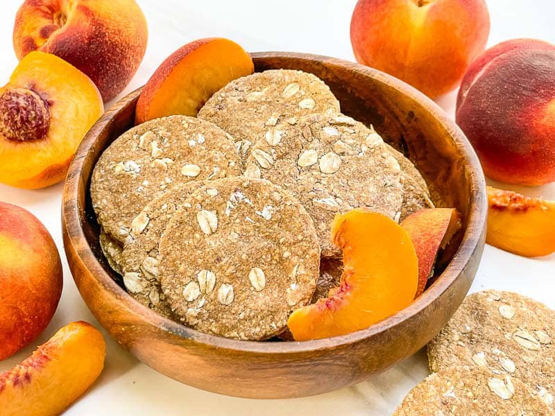 Peach Crisp Dog Treats in a wood bowl surrounded by fresh peaches.