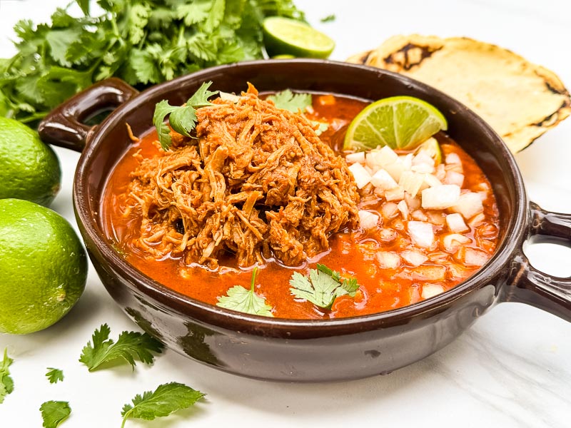 A brown round dish full of Chicken Birria (Birria de Pollo) and broth on a counter.