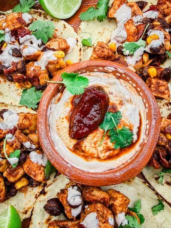 Looking down on Homemade Chipotle Crema Sauce in a small dish in the middle of tacos with fresh cilantro and a smoked chipotle pepper on top.