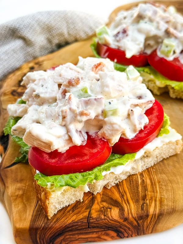 A pile of Bacon Ranch Chicken Salad sitting on top of tomatoes and lettuce on a slice of bread, on a cutting board.