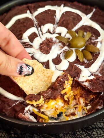 A hand digging a chip into Halloween Spider Bean Dip.