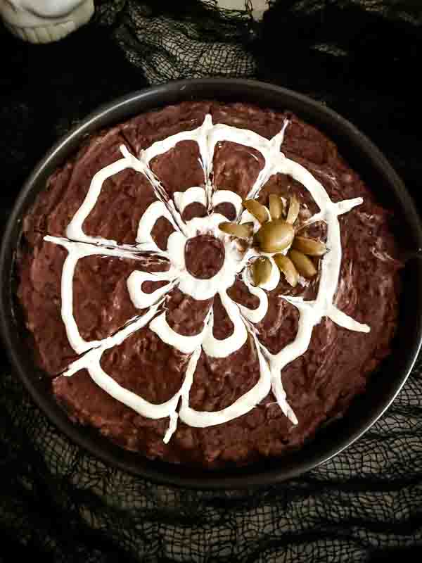 Looking down on a Halloween Spider Bean Dip with a white sour cream web design on a black bean dip and an olive spider.
