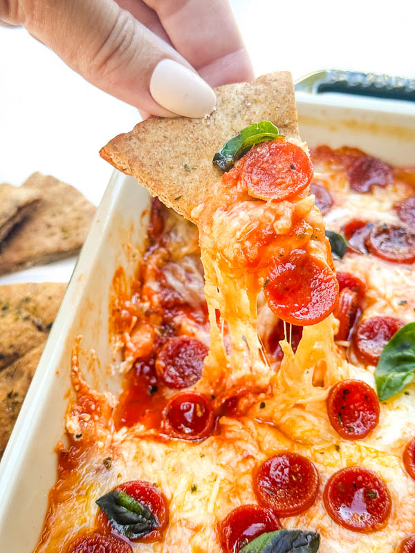 A hand dunking a pita chip into a cheesy Pepperoni Pizza Dip.